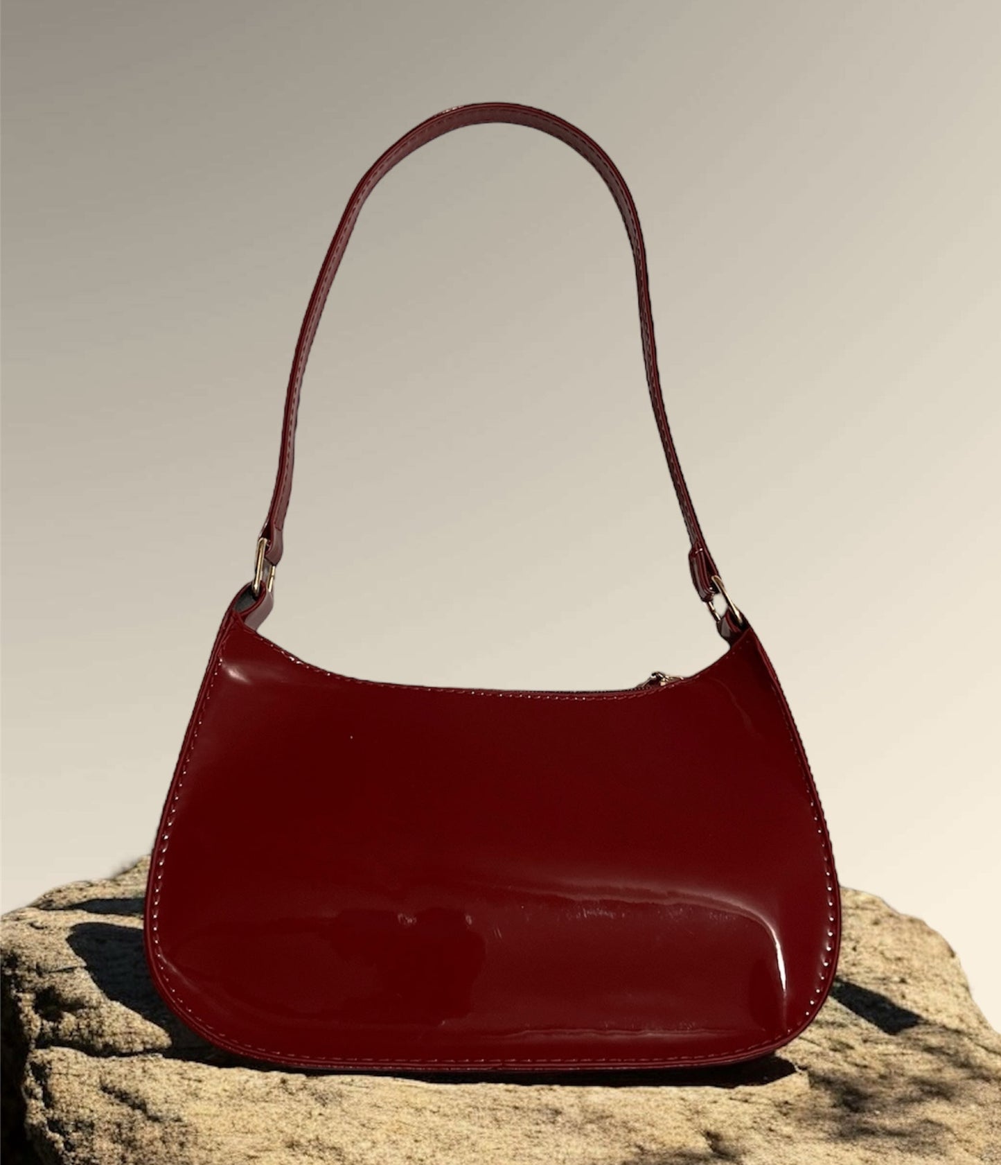 A maroon patent leather shoulder bag displayed on a rocky surface, showcasing a sleek design with a glossy finish.