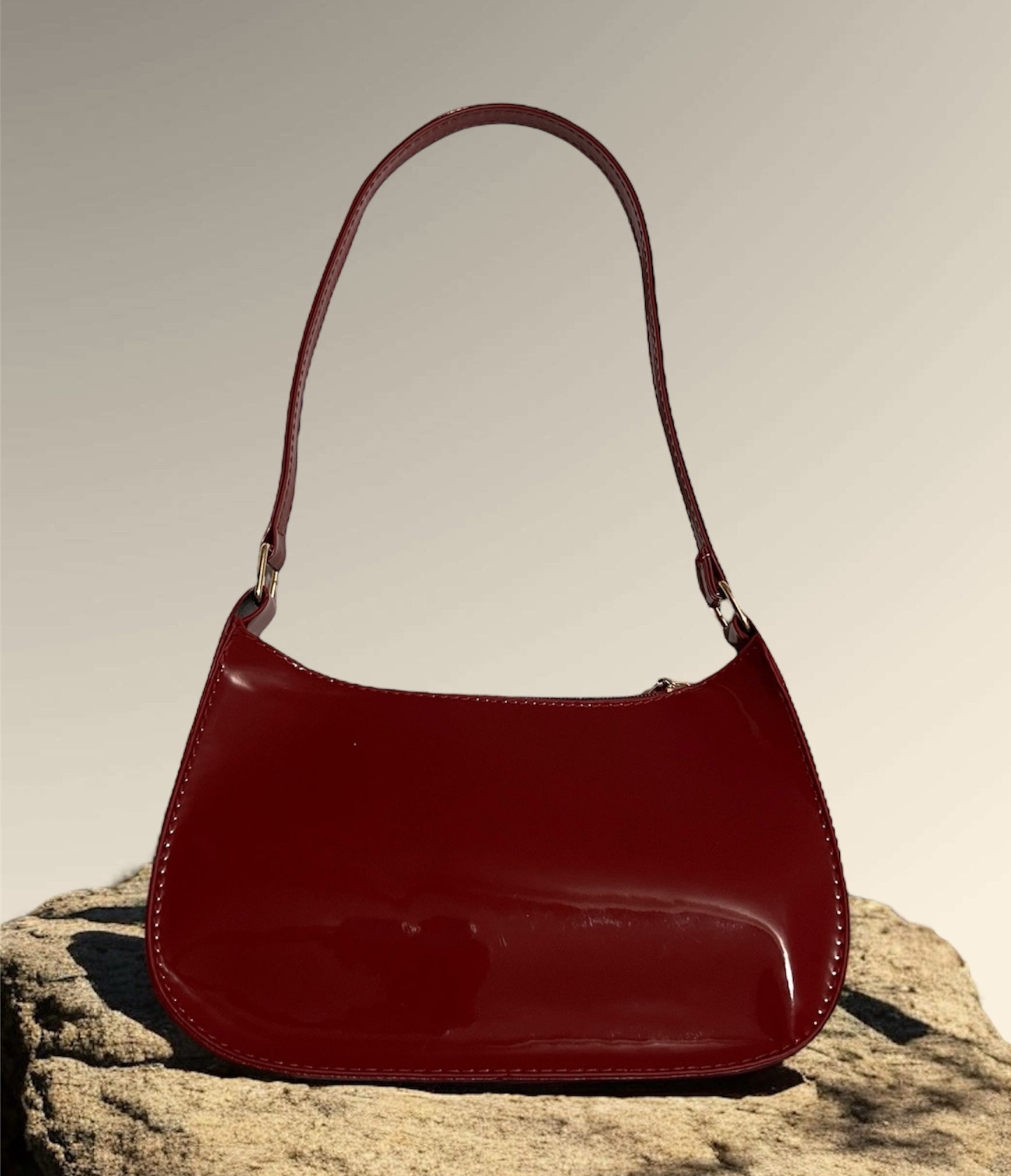 A maroon patent leather shoulder bag displayed on a rocky surface, showcasing a sleek design with a glossy finish.